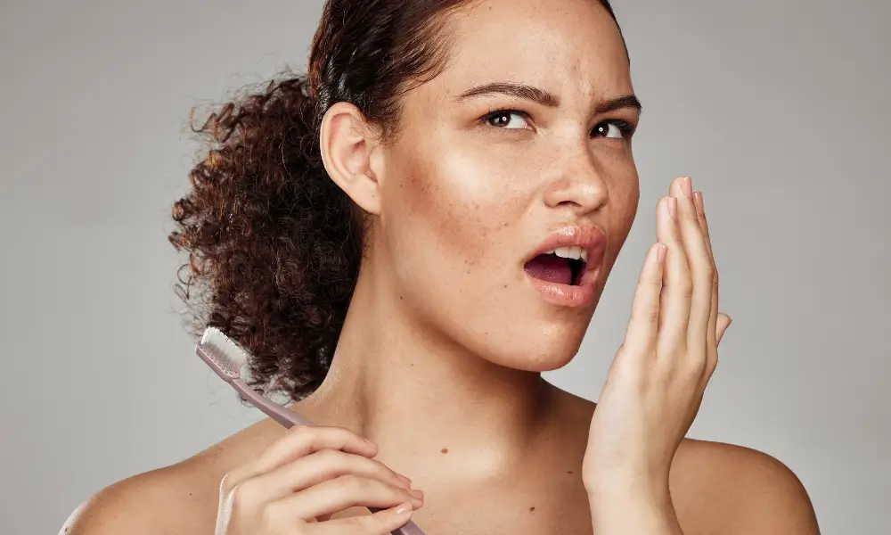 Woman holding a toothbrush and holding her other hand up to check her breath.
